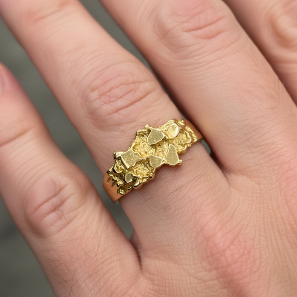 A close-up of a hand wearing a textured, organic-shaped gold ring with a black accent on one side, set against a blurred gray background.