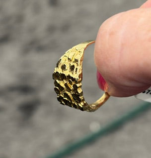 A close-up of a textured gold ring being held by a hand, with a blurred gray outdoor background.